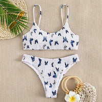 Bikini deux pièces texturé effet froissé imprimé papillons bleu à plat sur un tissu beige, avec chapeau de paille et fleur