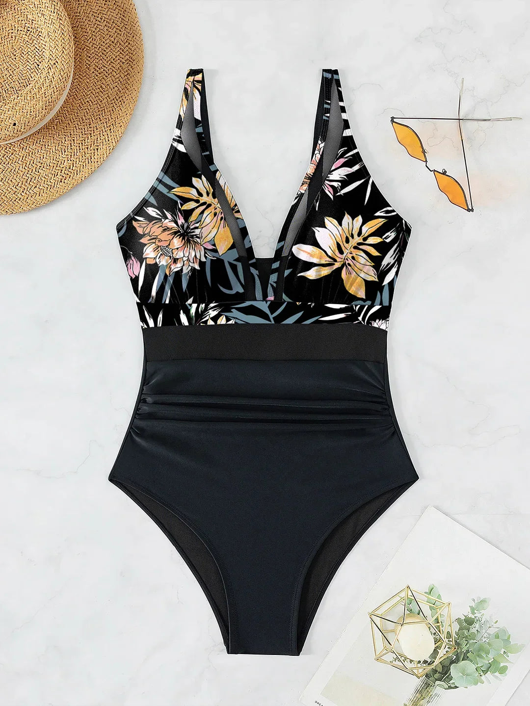 Maillot une pièce imprimé feuilles posé à plat sur marbre, haut à fleurs jaunes et feuilles grises et bas noir uni, chapeau en décor