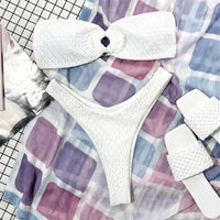 Bikini bandeau échancré blanc texturé posé à plat sur un tissu à carreaux, sandales blanches tressées à côté