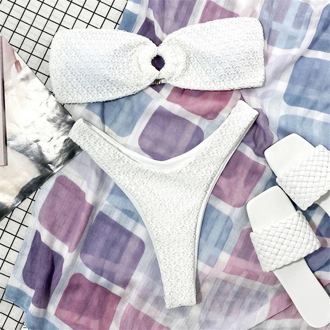 Bikini bandeau froncé blanc posé à plat sur un tissu aux carreaux violets et bleus, à côté de sandales tressées