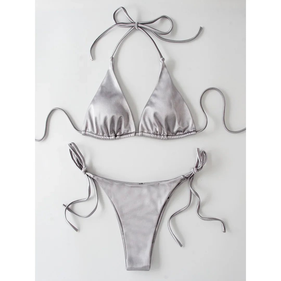 Bikini brésilien satiné gris argenté posé à plat sur fond blanc, haut triangle à bretelles de cou et bas à lacets latéraux