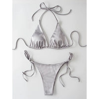 Bikini brésilien satiné gris argenté posé à plat sur fond blanc, haut triangle à bretelles de cou et bas à lacets latéraux