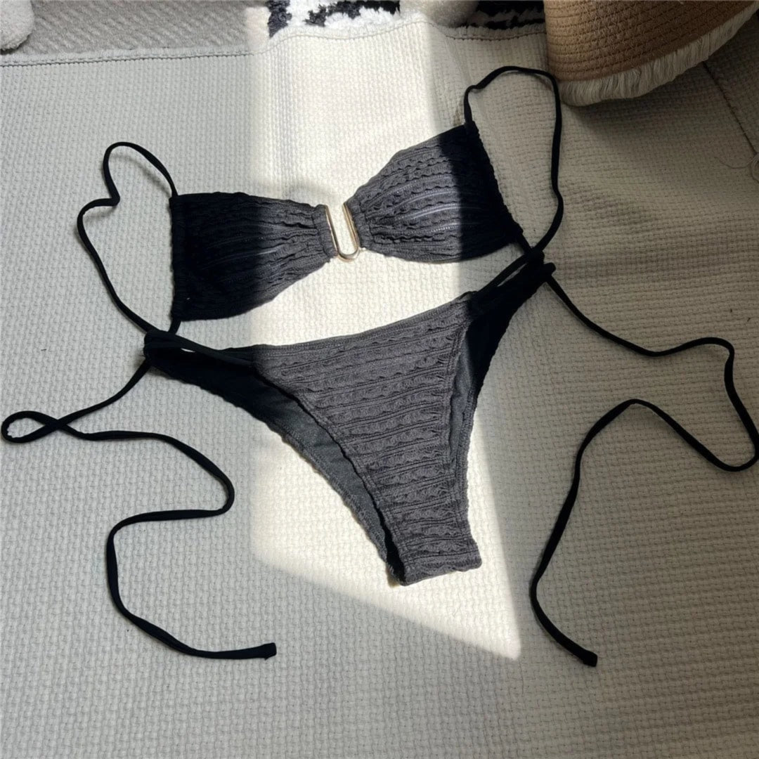 Bikini échancré texturé froissé noir étalé à plat sur un tapis gris au soleil, longues lanières et anneau doré au centre