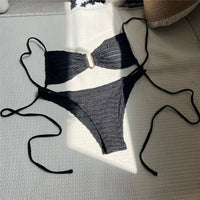Bikini échancré texturé froissé noir étalé à plat sur un tapis gris au soleil, longues lanières et anneau doré au centre