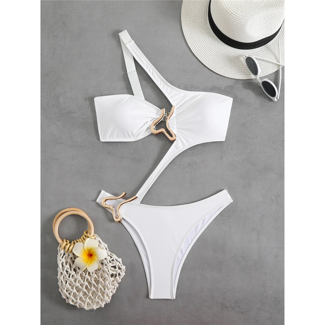 Maillot une pièce asymétrique blanc posé à plat sur fond gris, anneau doré au ventre, chapeau de paille et sac en filet