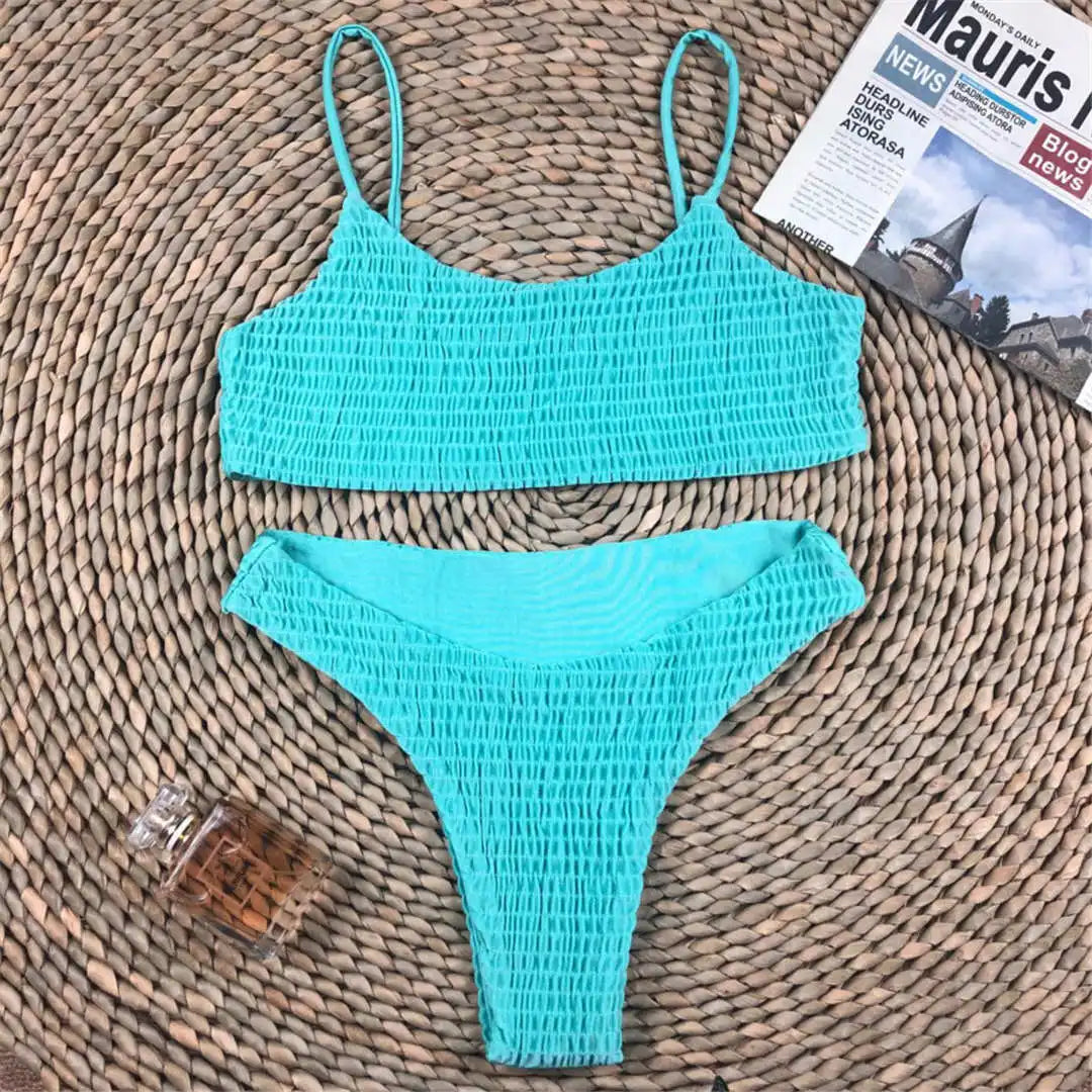 Bikini deux pièces texturé effet froissé turquoise à plat sur un tapis en osier, avec un magazine et un flacon