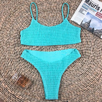 Bikini deux pièces texturé effet froissé turquoise à plat sur un tapis en osier, avec un magazine et un flacon