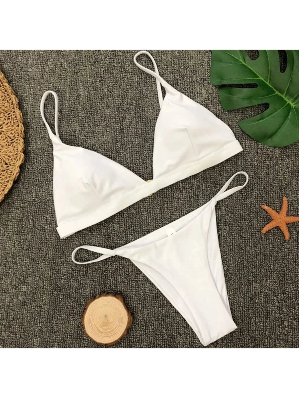 Bikini brésilien rembourré à taille basse blanc posé à plat sur tapis gris, rondin de bois et feuille de monstera