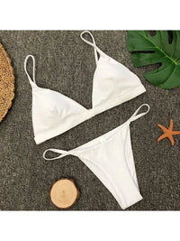 Bikini brésilien rembourré à taille basse blanc posé à plat sur tapis gris, rondin de bois et feuille de monstera
