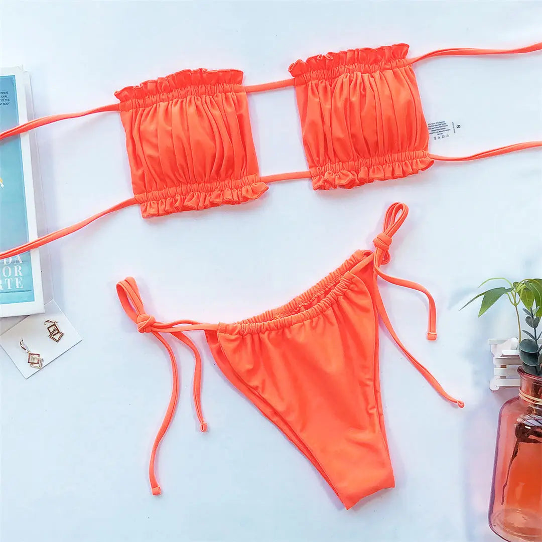 Bikini bandeau à volants bretelles réglables orange posé à plat sur fond bleu clair, avec un magazine