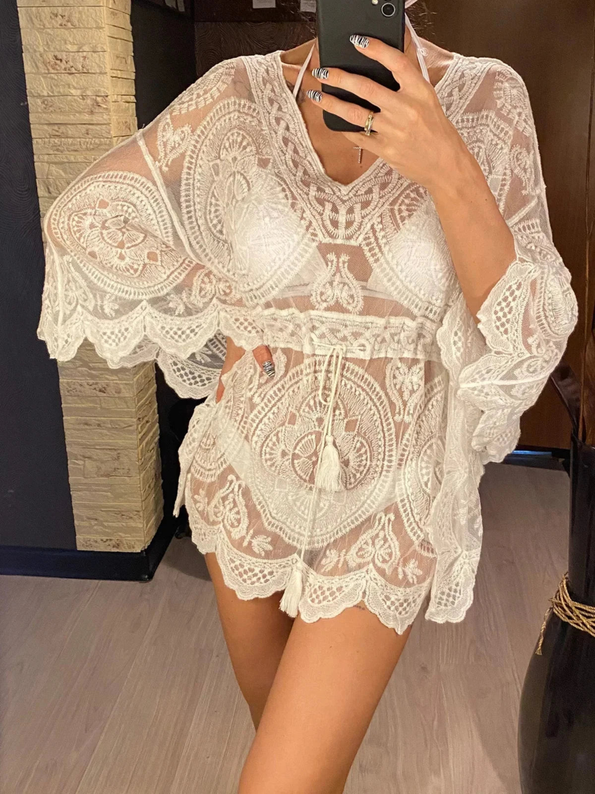 Tunique de plage blanche en dentelle à manches évasées portée de face par une femme qui se photographie, lien à la taille à pompons