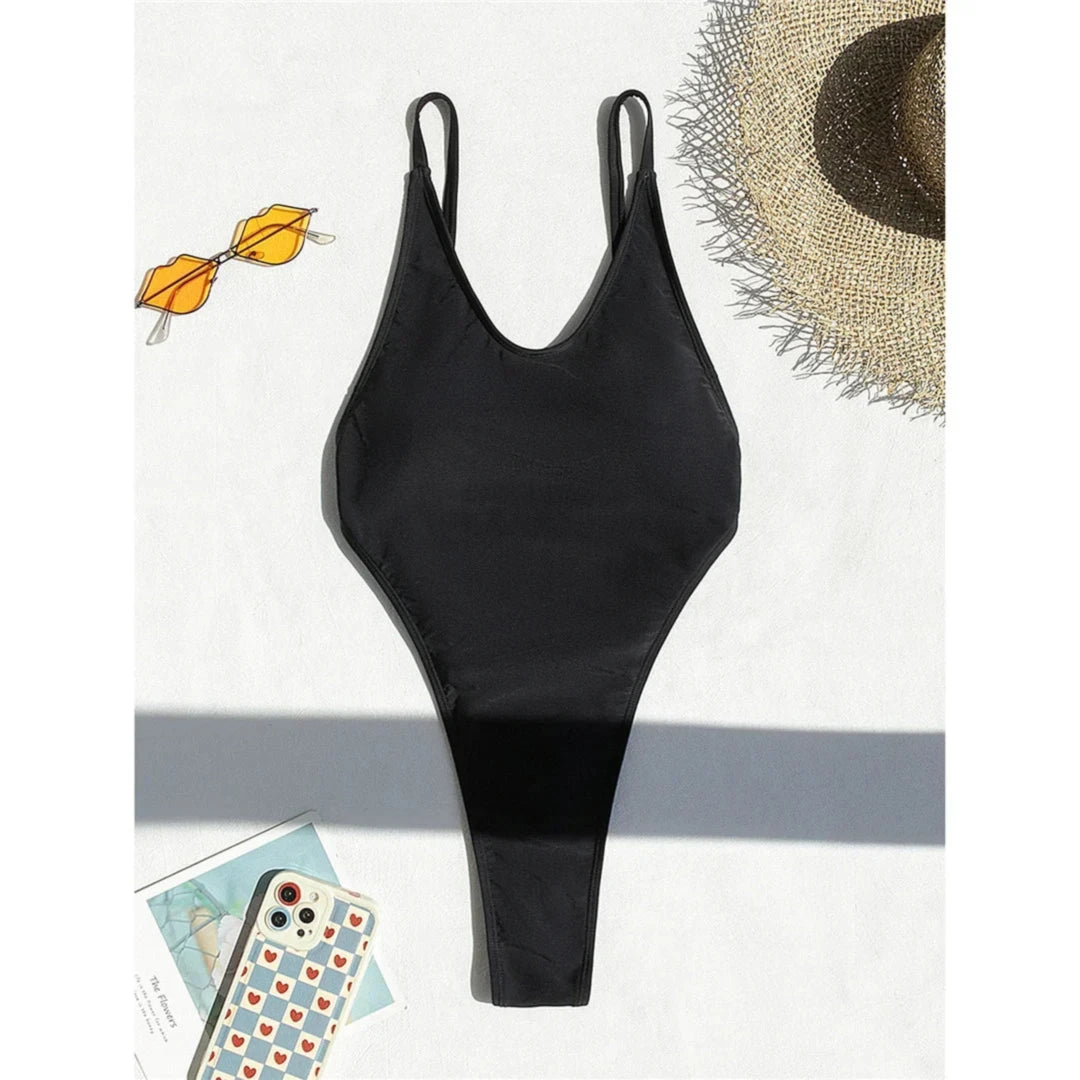 Maillot de bain une pièce noir posé à plat sur fond blanc, bretelles fines et échancrure haute, lunettes à côté
