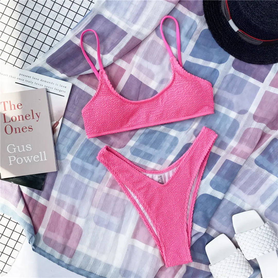 Bikini triangle rose vif effet froissé posé à plat sur tapis carreaux, à côté d'un chapeau noir et de sandales blanches