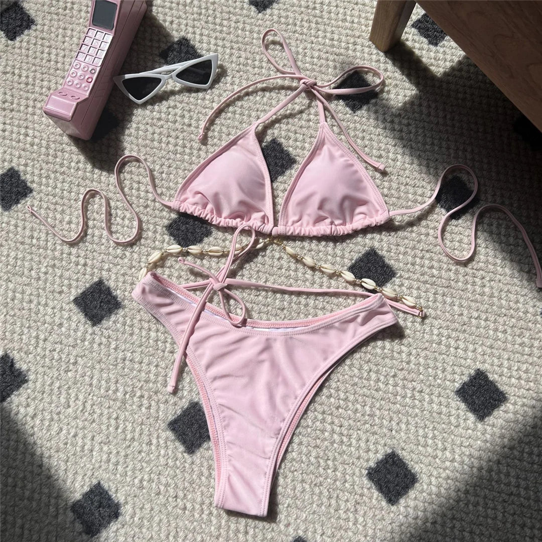 Bikini brésilien rose poudré posé à plat sur un tapis, triangle sans armatures et lanières ornées de coquilles dorées