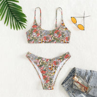 Bikini brésilien imprimé fleuri orange et vert posé à plat sur fond blanc, avec une feuille de palmier et des lunettes jaunes