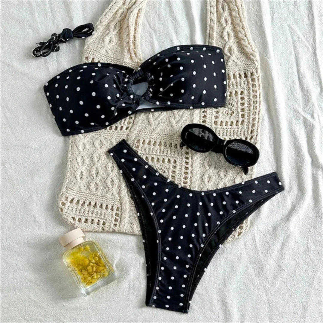 Bikini bandeau brésilien à pois noir posé à plat sur un tissu crochet crème, avec lunettes et flacon à côté