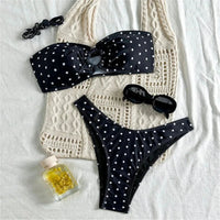Bikini bandeau brésilien à pois noir posé à plat sur un tissu crochet crème, avec lunettes et flacon à côté