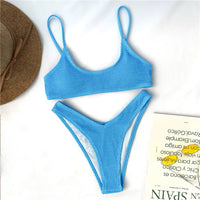 Bikini triangle effet froissé bleu posé à plat sur fond blanc, à côté d'un chapeau de paille et d'un magazine
