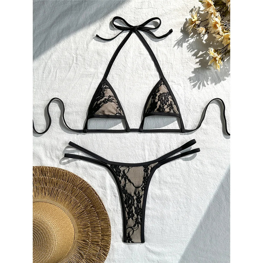 Bikini string micro noir en dentelle, bordure fine et bretelles croisées, posé à plat sur tissu blanc avec chapeau de paille