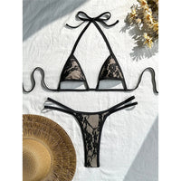 Bikini string micro noir en dentelle, bordure fine et bretelles croisées, posé à plat sur tissu blanc avec chapeau de paille
