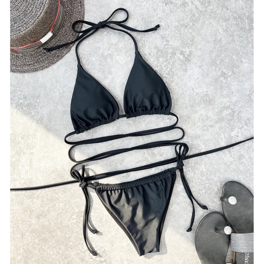 Bikini brésilien noir étalé à plat sur un béton gris avec un chapeau de paille et des sandales noires