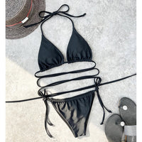 Bikini brésilien noir étalé à plat sur un béton gris avec un chapeau de paille et des sandales noires