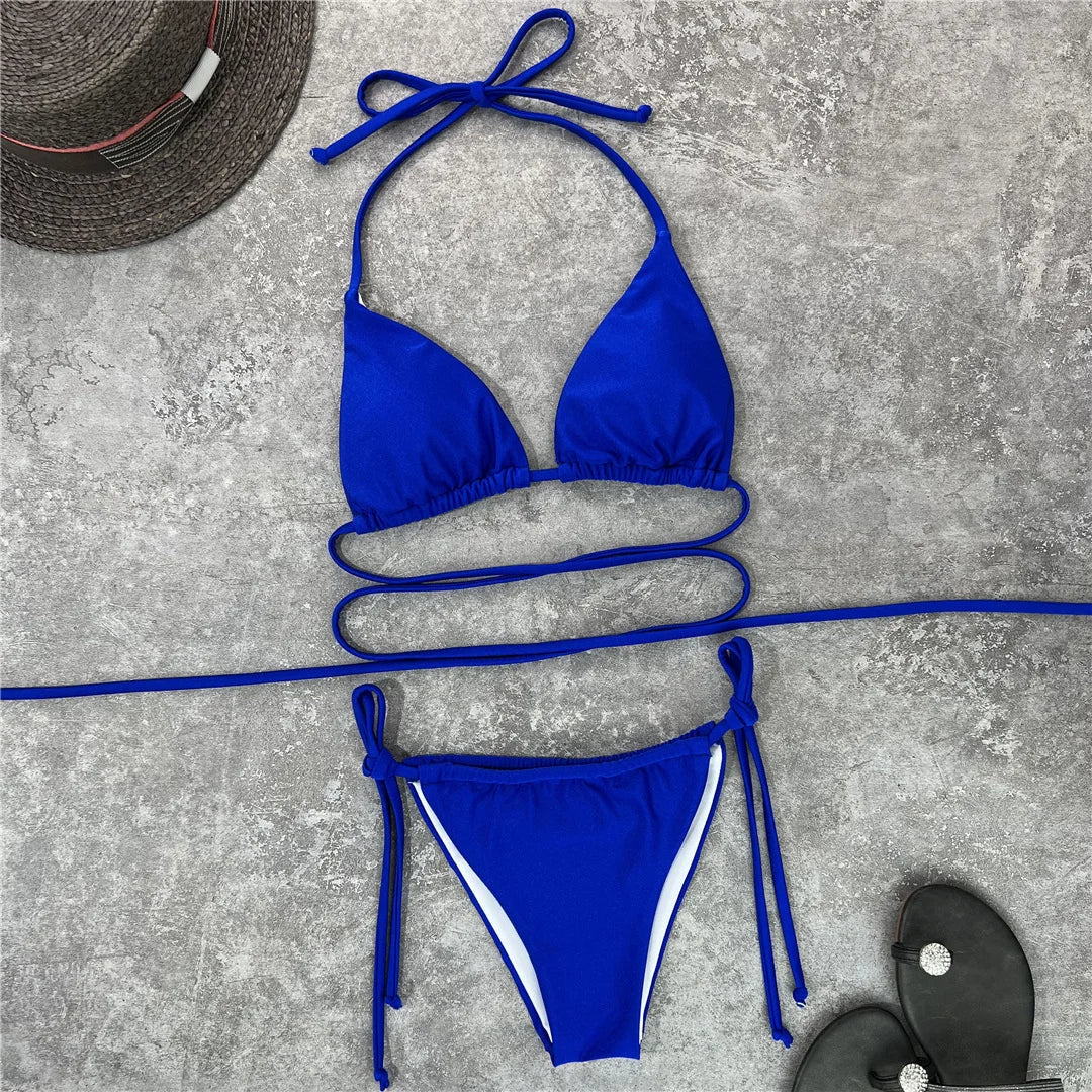 Bikini brésilien bleu roi étalé à plat sur un béton gris avec un chapeau de paille et des sandales noires
