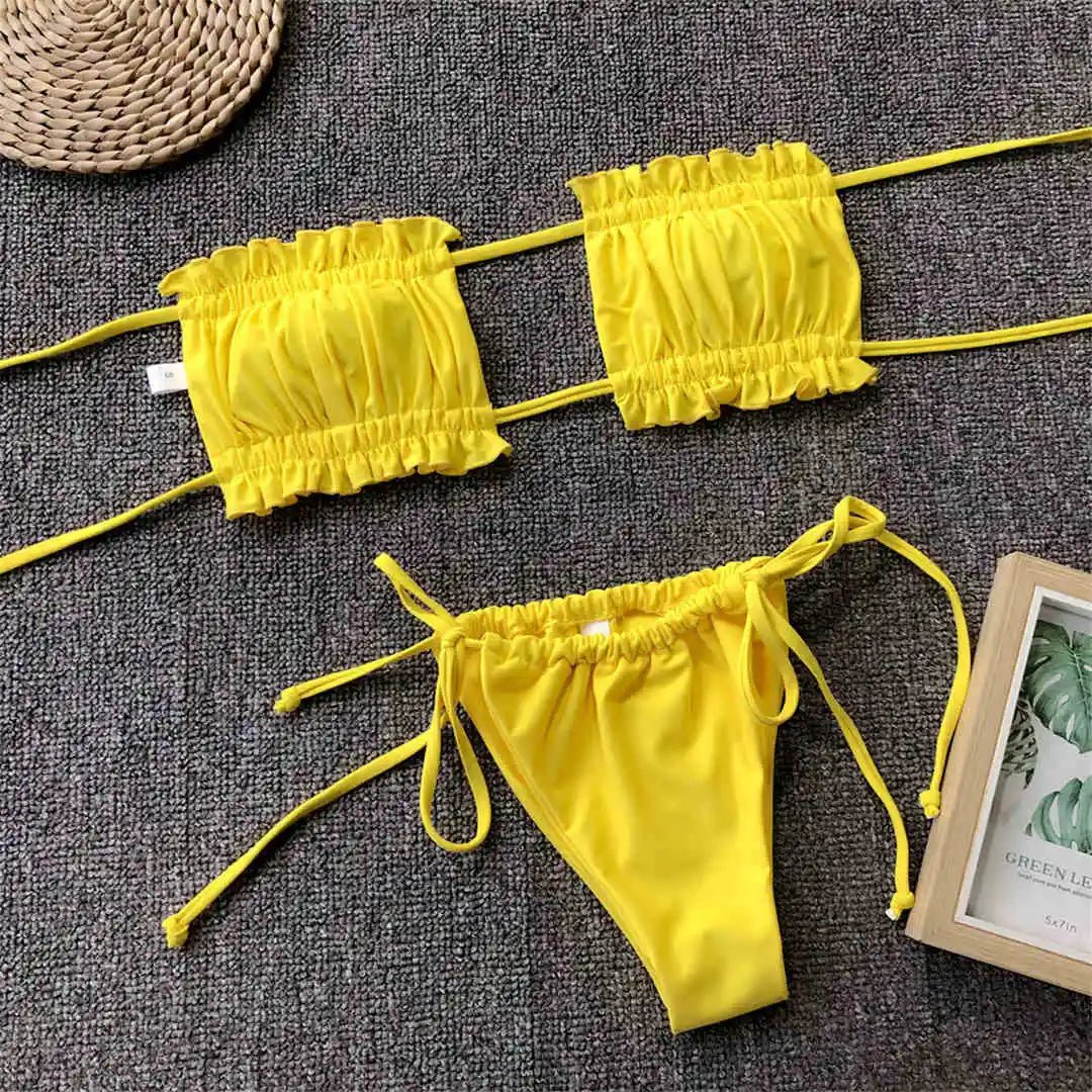 Bikini jaune à bandeau froncé posé à plat sur un tapis gris, à côté d'un magazine, bas à liens noués sur les côtés