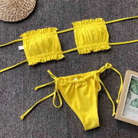 Bikini bandeau à volants bretelles réglables jaune posé à plat sur un tapis gris, à côté d'un magazine