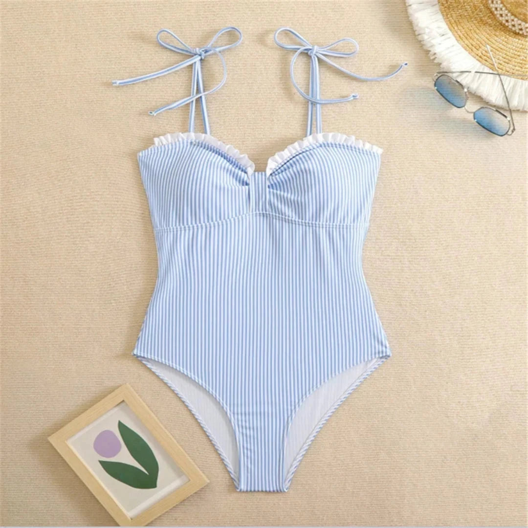 Maillot une pièce à volants rayé bleu et blanc posé à plat sur du sable beige, avec un chapeau de paille et un cadre