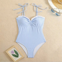 Maillot une pièce à volants rayé bleu et blanc posé à plat sur du sable beige, avec un chapeau de paille et un cadre