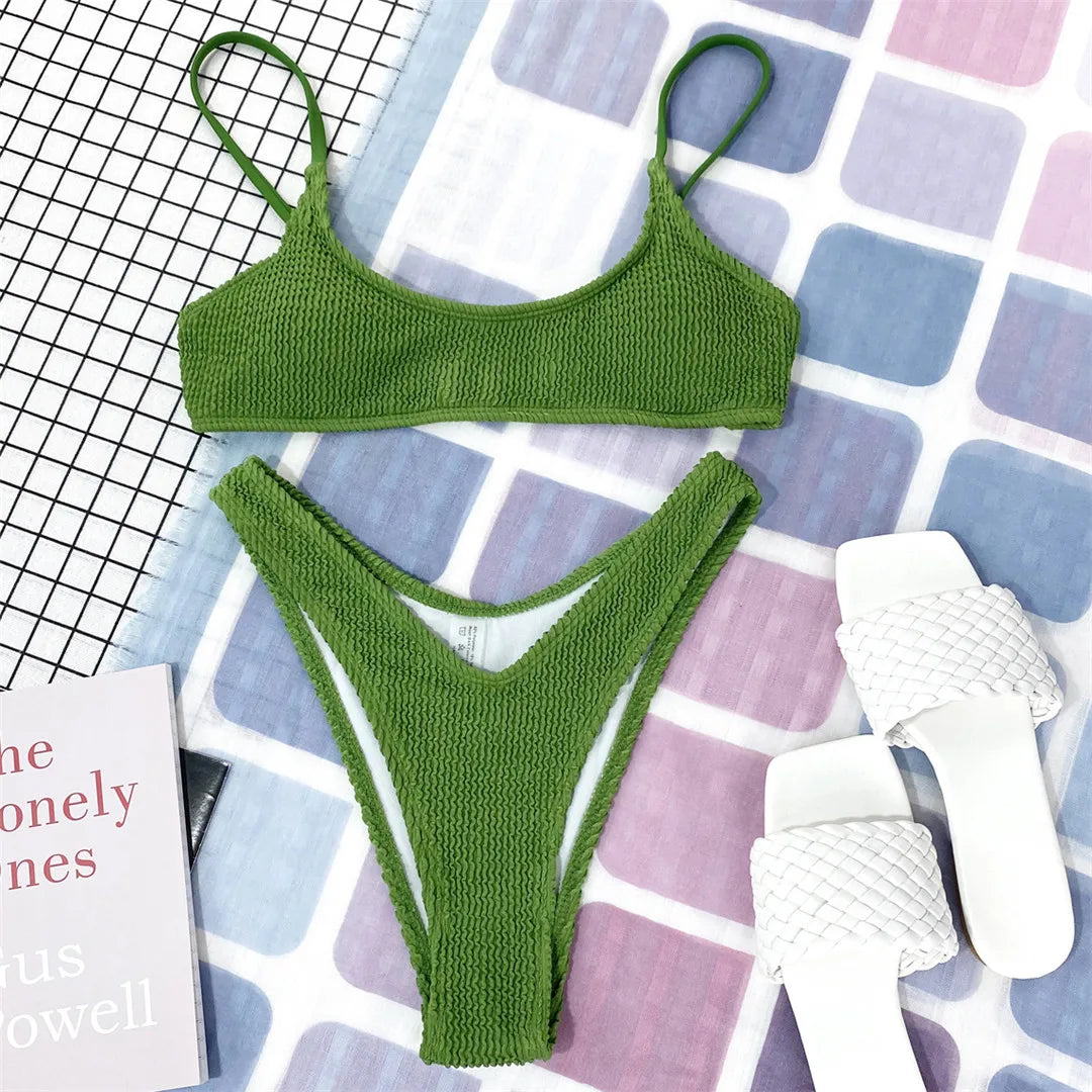 Bikini triangle effet froissé vert kaki posé à plat sur un tapis carreaux, à côté de sandales blanches et d'un livre