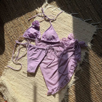 Maillot violet clair à plat sur un tapis en fibres, haut à fleur en relief et bas à liens, pareo volanté posé à côté