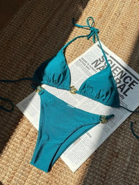 Bikini tour de cou à anneau métallique bleu paon posé à plat sur un journal, liens fins et anneaux dorés sur le haut et le bas