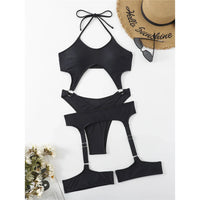 Maillot de bain noir à découpes et sangles de jarretière posé à plat sur fond blanc, chapeau de paille et fleurs à côté