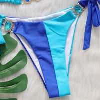 Gros plan sur le bas bleu et bleu clair du bikini patchwork, anneau ovale orné de strass turquoise sur la hanche et nœud latéral