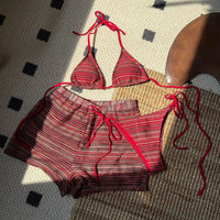 Maillot trois pièces tour de cou rayé rouge à plat sur un tapis, lanières rouges nouées et short à rayures rouges