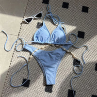 Bikini brésilien bleu clair posé à plat sur un tapis, triangle sans armatures et lien doré en coquilles sur le bas