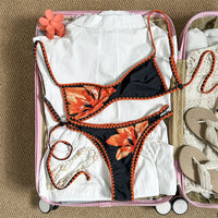 Bikini brésilien triangle imprimé fleuri noir et orange à plat dans une valise, fleurs orange et bordure au crochet orange