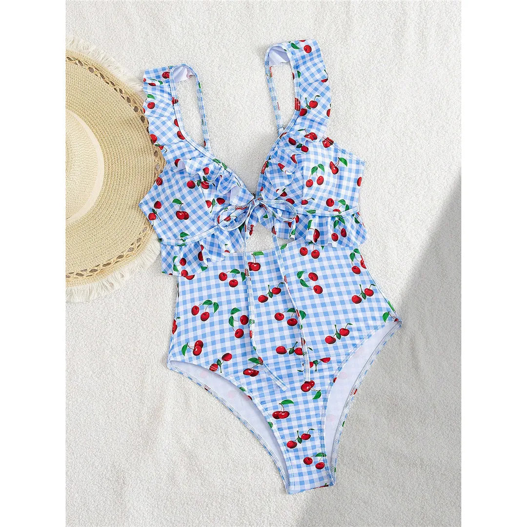 Maillot de bain une pièce échancré bleu clair à cerises posé à plat sur fond blanc, bretelles fines à volants