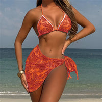 Maillot trois pièces rouge orangé porté de face par une femme debout sur la plage, paréo noué sur la hanche