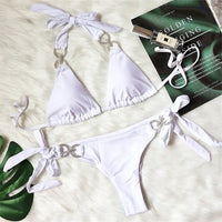 Bikini brésilien tour de cou à ornements strass blanc posé à plat sur une fourrure blanche, près d'un magazine et de flacons