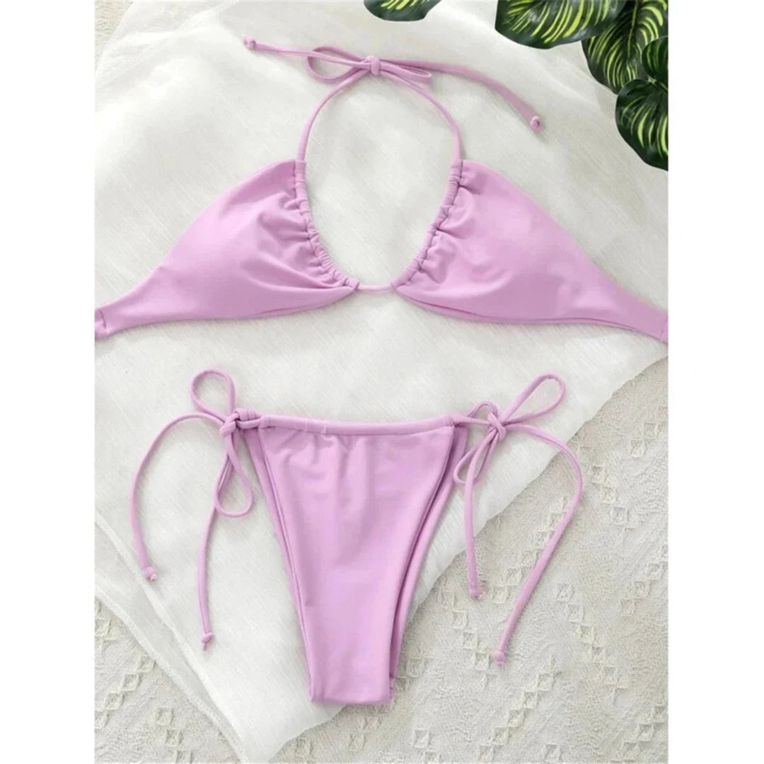 Bikini string mini tour de cou taille basse violet clair posé à plat sur un tissu blanc, feuille verte en haut de l'image