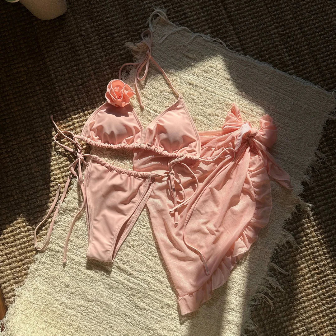 Maillot trois pièces rose pêche posé à plat sur un tapis en fibres, bas à liens et pareo volanté posé à côté