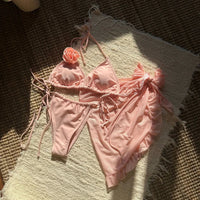 Maillot trois pièces rose pêche posé à plat sur un tapis en fibres, bas à liens et pareo volanté posé à côté