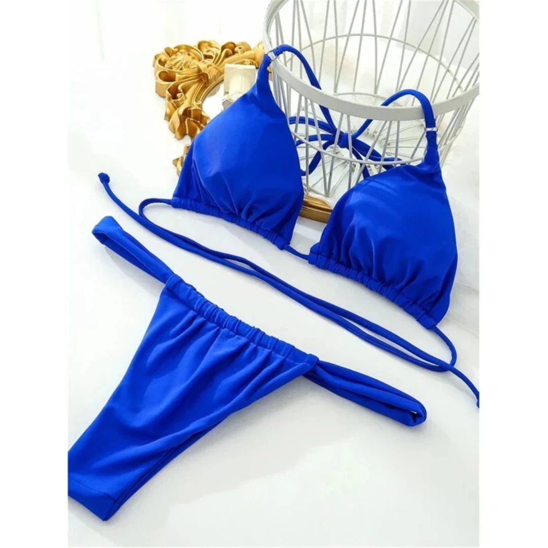 Bikini string bleu roi posé à plat sur un fond blanc près d'un panier en métal, liens croisés au dos du haut